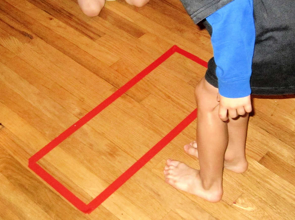 Child standing next to a red tape rectangle on the floor.