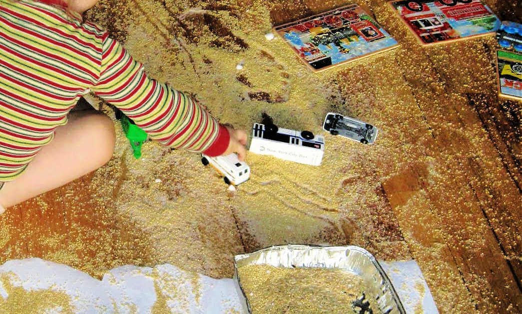 Child playing with cars and making a mess on the floor with a tray of millet