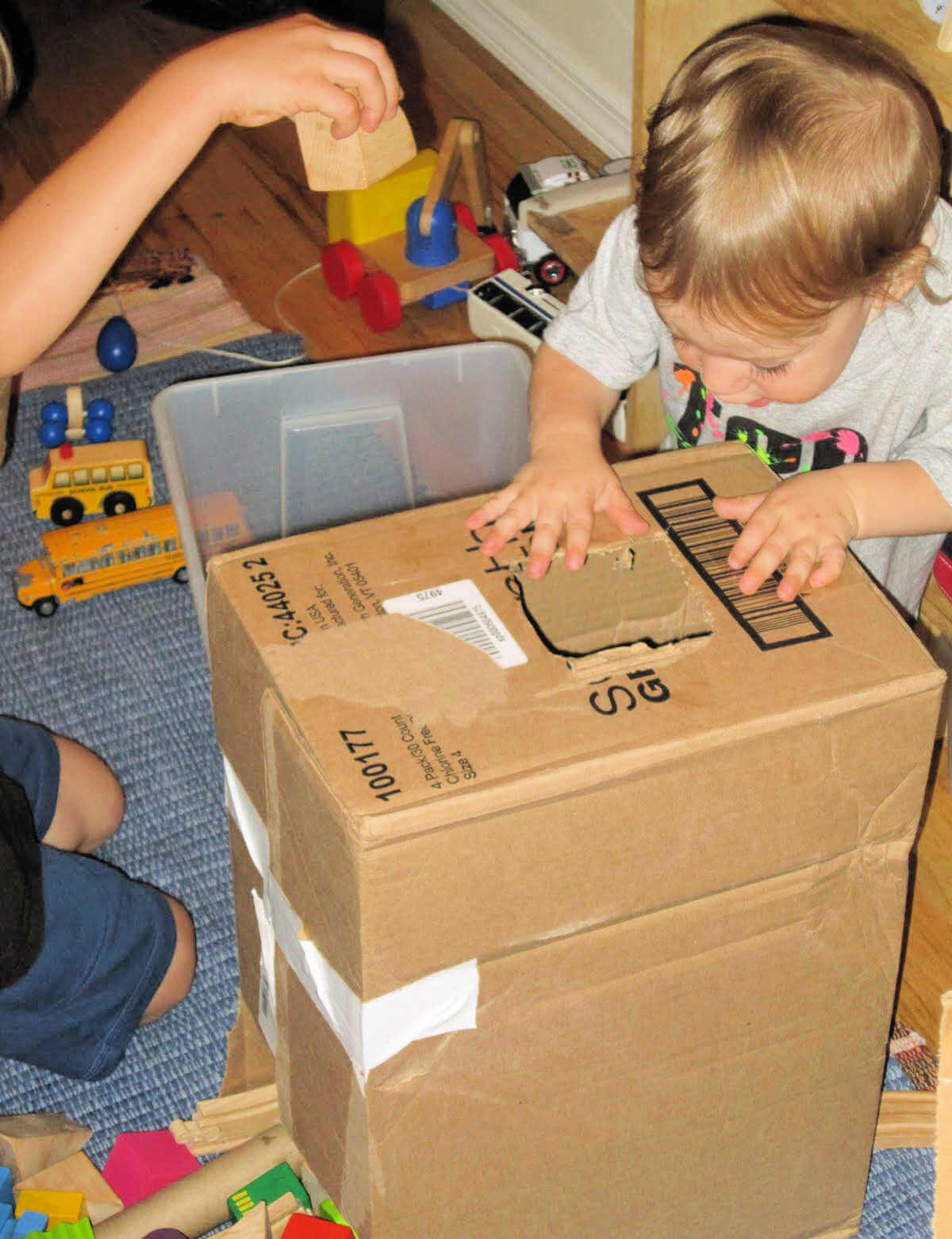 Two children dropping toy blocks into a hole cut in a cardboard box